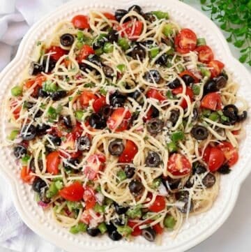 A white plate filled with spaghetti pasta salad, topped with black olives, cherry tomatoes, chopped green bell peppers, and shredded cheese. The colorful salad is presented on a textured white cloth.