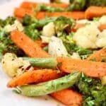 A close-up of roasted vegetables, including carrots, broccoli, cauliflower, and green beans, seasoned with herbs and served on a white plate.