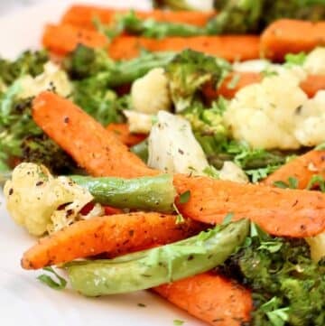 A close-up of roasted vegetables, including carrots, broccoli, cauliflower, and green beans, seasoned with herbs and served on a white plate.