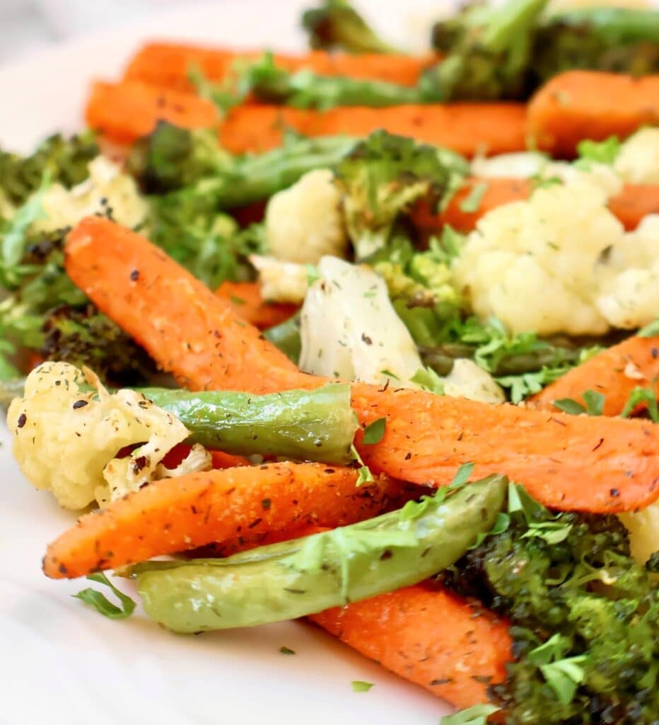 A close-up of roasted vegetables, including carrots, broccoli, cauliflower, and green beans, seasoned with herbs and served on a white plate.