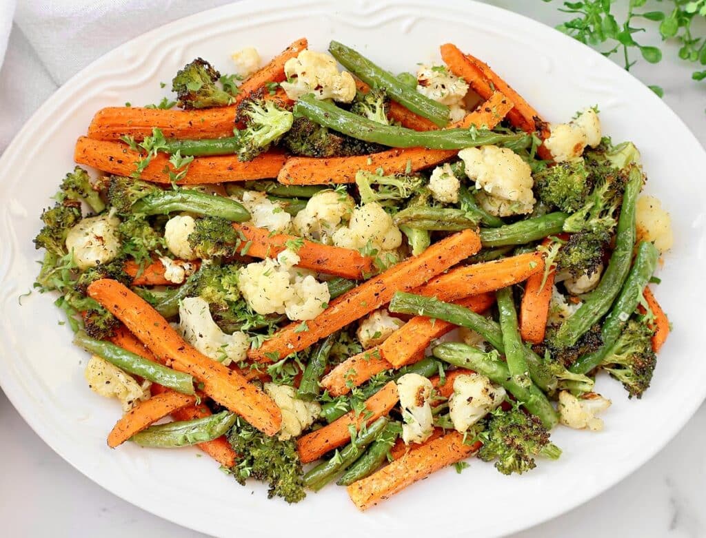 A white plate filled with roasted vegetables, including carrots, green beans, broccoli, and cauliflower, garnished with fresh herbs.