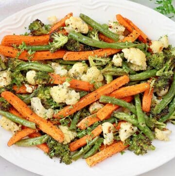 A white plate filled with roasted vegetables, including carrots, green beans, broccoli, and cauliflower, garnished with fresh herbs.