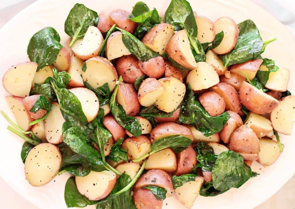 A platter of roasted red potatoes mixed with fresh spinach leaves, seasoned with black pepper and herbs, served on a white dish.