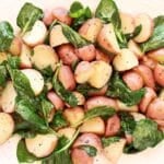 A platter of roasted red potatoes mixed with fresh spinach leaves, seasoned with black pepper and herbs, served on a white dish.