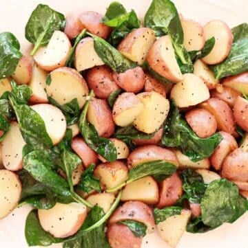 A platter of roasted red potatoes mixed with fresh spinach leaves, seasoned with black pepper and herbs, served on a white dish.