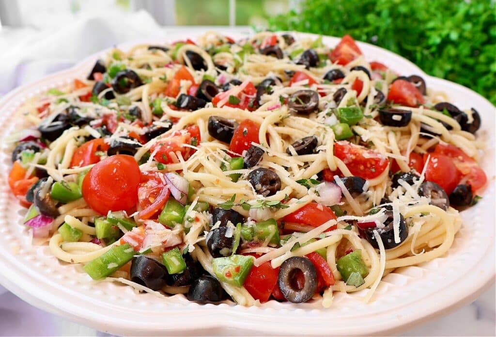 A plate of spaghetti salad topped with sliced black olives, cherry tomatoes, chopped green bell peppers, red onions, shredded cheese, and herbs. The dish appears fresh and colorful with visible vegetables.