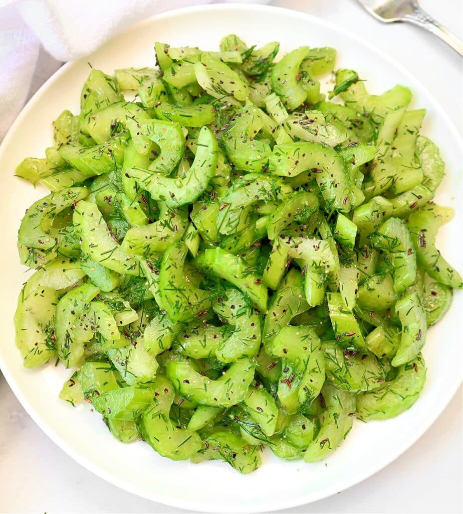 A white plate filled with sliced cucumber salad garnished with chopped fresh dill and black pepper, placed on a light surface.