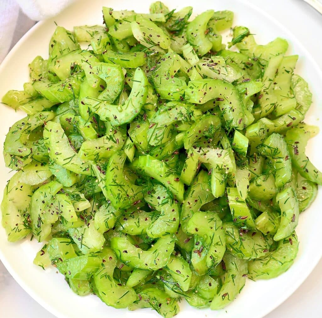 A white plate filled with sliced celery salad, garnished with fresh dill and sprinkled with black seeds or spices. The salad appears fresh and vibrant green.