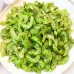 A white plate filled with sliced celery salad, garnished with fresh dill and sprinkled with black seeds or spices. The salad appears fresh and vibrant green.