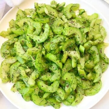 A white plate filled with sliced celery salad, garnished with fresh dill and sprinkled with black seeds or spices. The salad appears fresh and vibrant green.