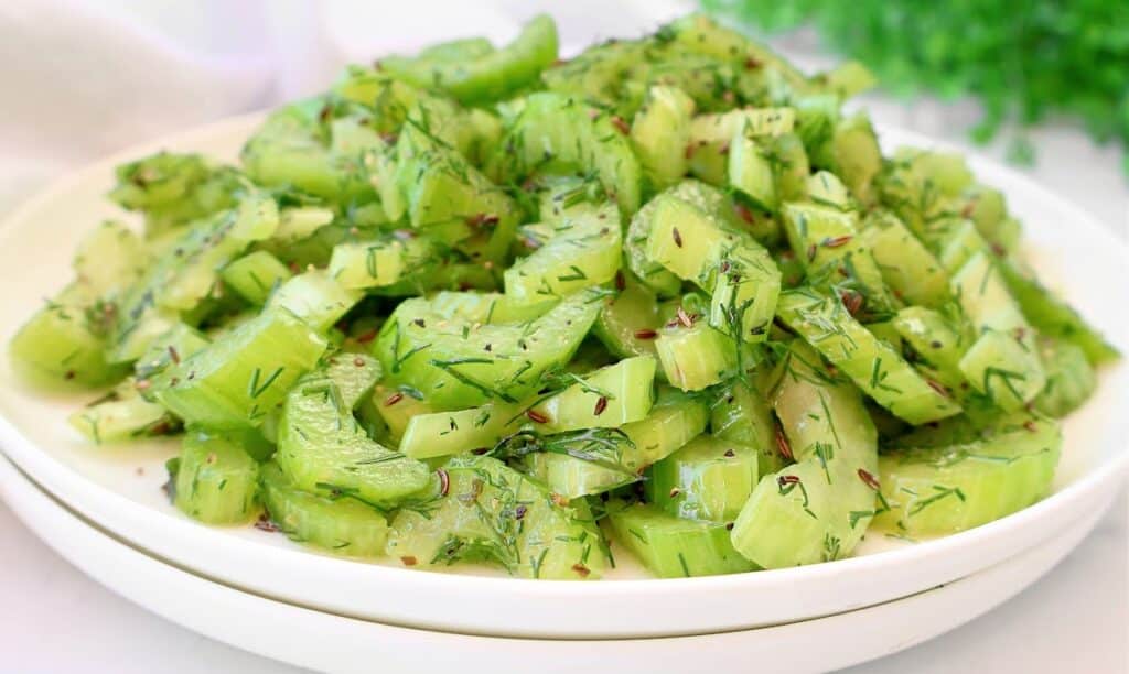 A white plate filled with sliced celery mixed with fresh dill and spices, set on another white plate with a blurred green background.