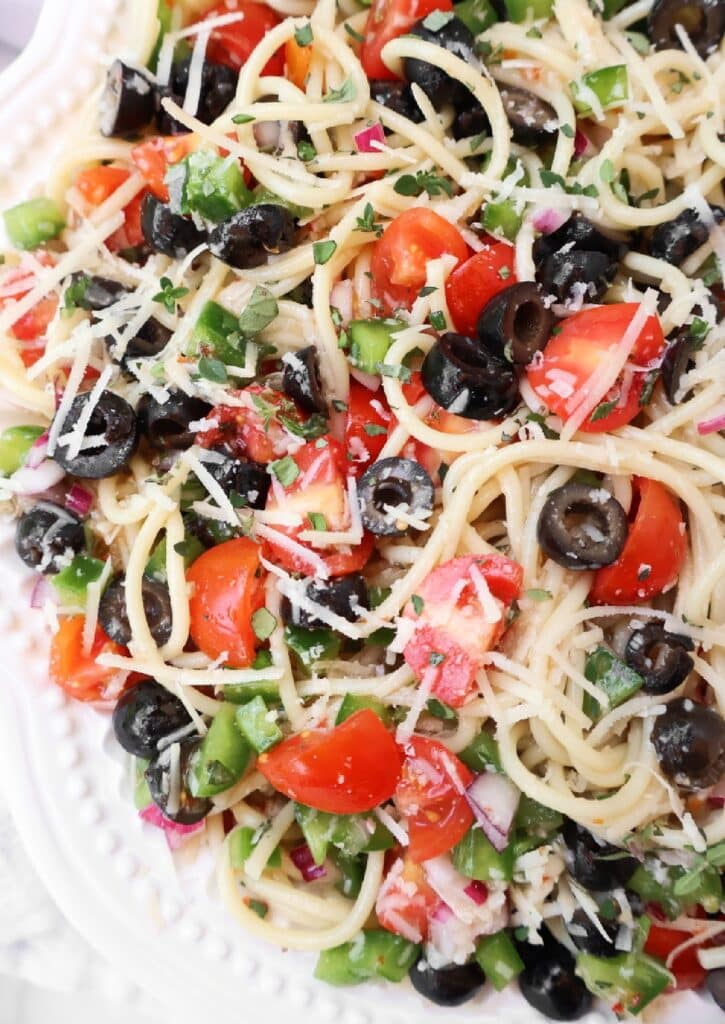 A close-up of a pasta salad with spaghetti, black olives, cherry tomatoes, green bell peppers, red onions, and grated Parmesan cheese on a white plate.