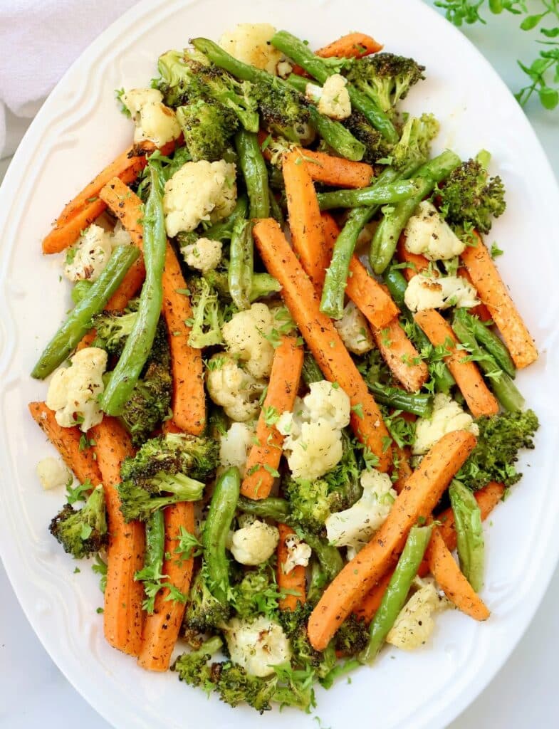 A white platter filled with roasted vegetables including carrot sticks, green beans, broccoli, and cauliflower, garnished with chopped herbs.