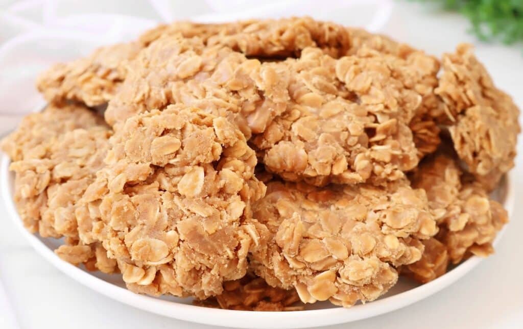 A plate stacked with several no-bake cookies made with oats and a light brown, creamy mixture, likely peanut butter, sitting on a white surface.