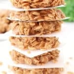 A stack of round, chewy oatmeal cookies separated by pieces of white parchment paper, with a blurred green and white background.