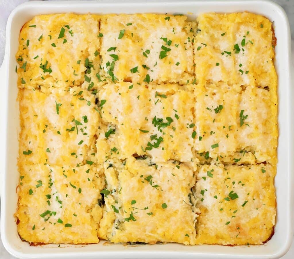 A baked casserole in a white dish, cut into twelve square pieces and garnished with chopped parsley on top. The casserole appears creamy with a golden, slightly browned surface.