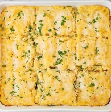 A baked casserole in a white dish, cut into twelve square pieces and garnished with chopped parsley on top. The casserole appears creamy with a golden, slightly browned surface.