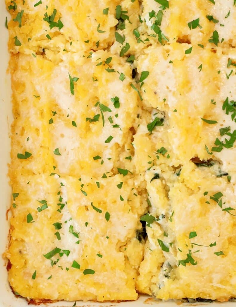 A close-up of a baked casserole cut into rectangular pieces, topped with melted cheese and sprinkled with chopped fresh parsley.
