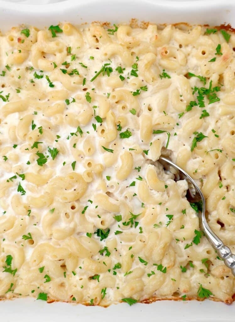 A close-up of make ahead mac and cheese in a white dish, topped with chopped parsley. A serving spoon is scooping out a portion from the corner.