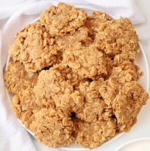 A white plate piled with homemade no-bake cookies made from oats and a light brown peanut butter mixture, set on a white surface with a white cloth nearby.
