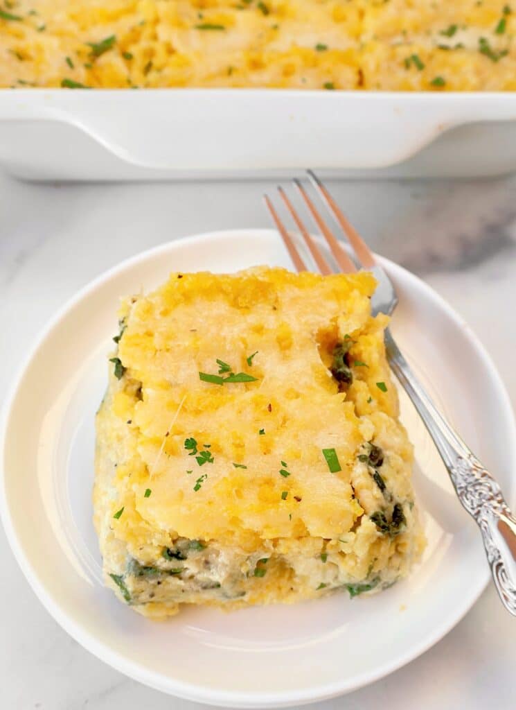 A serving of cheesy casserole with spinach on a white plate and a fork, garnished with chopped herbs, with the casserole dish in the background.