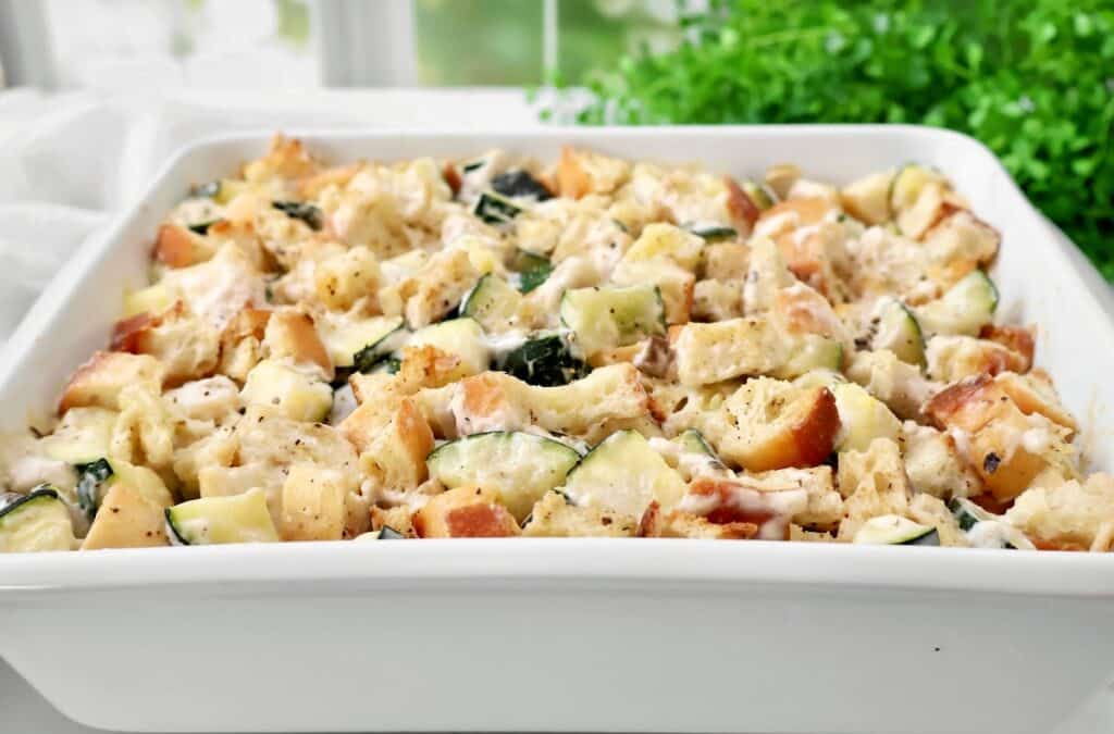 A white casserole dish filled with a baked bread and vegetable casserole, featuring chunks of bread, zucchini, and melted cheese, with a green plant and a bright window in the background.