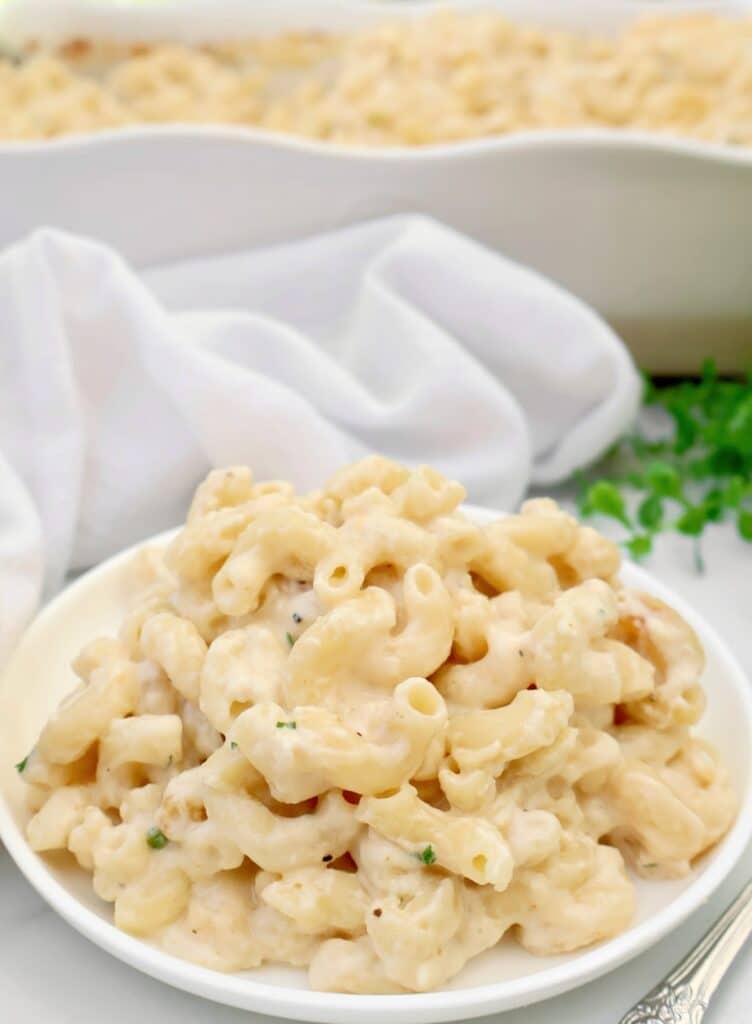 A plate of creamy make ahead mac and cheese is served in front of a white baking dish. The macaroni is topped with a rich cheese sauce and garnished with herbs, with a white napkin and greenery in the background.