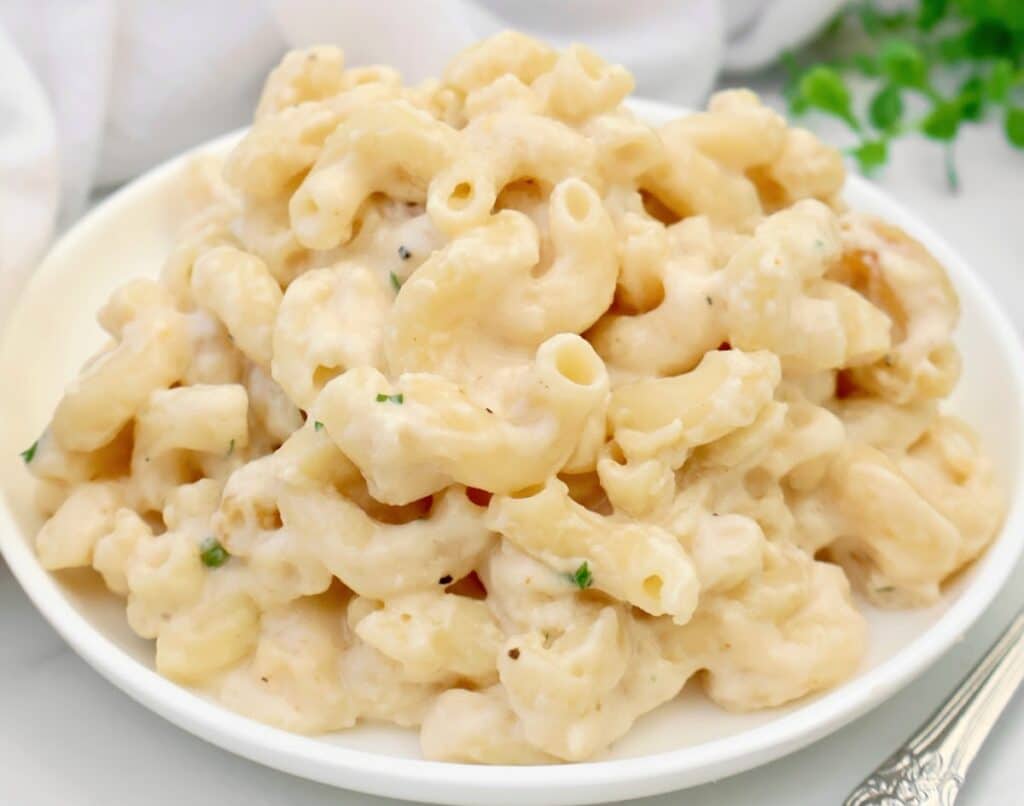 A white plate filled with creamy make ahead mac and cheese, made with elbow pasta and a rich cheese sauce, garnished with a few small green herbs.