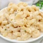 A white plate filled with creamy make ahead mac and cheese, made with elbow pasta and a rich cheese sauce, garnished with a few small green herbs.
