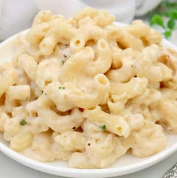 A white plate filled with creamy make ahead mac and cheese, made with elbow pasta and a rich cheese sauce, garnished with a few small green herbs.