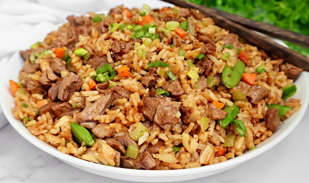 A bowl of vegan beef steak fried rice with chopped carrots, green beans, and scallions, garnished with black pepper. Chopsticks rest on the edge of the bowl.