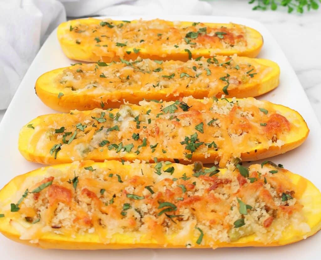 Four halves of stuffed yellow squash are topped with melted cheese, breadcrumbs, and chopped herbs, arranged on a white rectangular plate. The golden dish looks perfectly garnished and is set on a light surface.