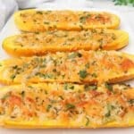 Four halves of stuffed yellow squash are topped with melted cheese, breadcrumbs, and chopped herbs, arranged on a white rectangular plate. The golden dish looks perfectly garnished and is set on a light surface.