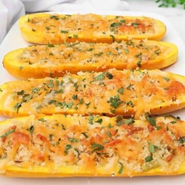 Four halves of stuffed yellow squash are topped with melted cheese, breadcrumbs, and chopped herbs, arranged on a white rectangular plate. The golden dish looks perfectly garnished and is set on a light surface.