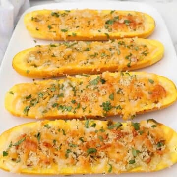 Four stuffed yellow squash halves topped with melted cheese, breadcrumbs, and chopped herbs are arranged on a white rectangular plate.