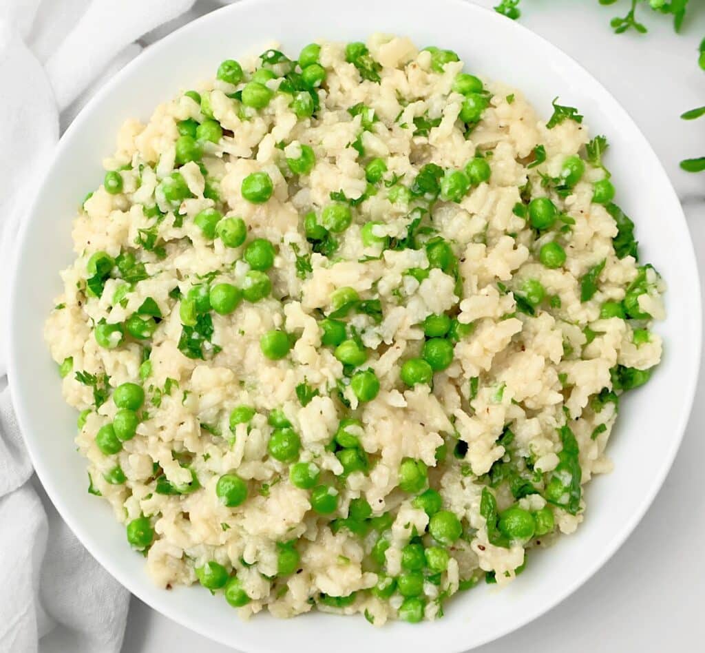 A white bowl filled with creamy slow cooker risotto mixed with green peas and garnished with chopped herbs, placed on a white surface with a white napkin nearby.