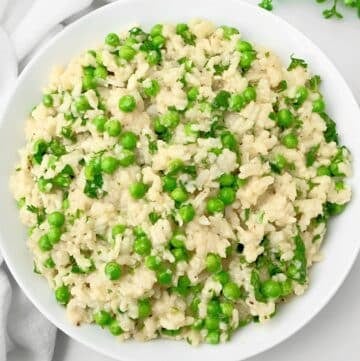 A white bowl filled with creamy slow cooker risotto mixed with green peas and garnished with chopped herbs, placed on a white surface with a white napkin nearby.