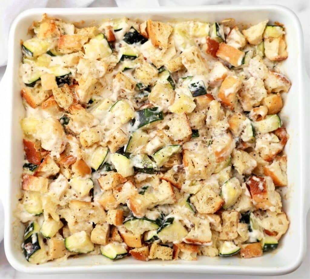 A white baking dish filled with a casserole made of cubed bread, zucchini pieces, melted cheese, and herbs, all mixed and baked to a golden brown.