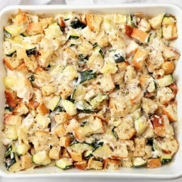 A white baking dish filled with a casserole made of cubed bread, zucchini pieces, melted cheese, and herbs, all mixed and baked to a golden brown.