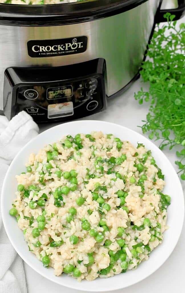 A white bowl filled with creamy slow cooker risotto and green peas sits in front of a Crock-Pot, with fresh herbs and a white napkin nearby.
