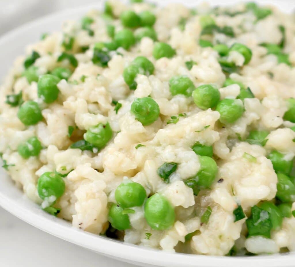 Creamy slow cooker risotto with bright green peas and fresh herbs, served on a white plate. The rice appears tender and slightly glossy, with peas evenly distributed throughout.
