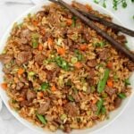 A bowl of vegan steak fried rice features hearty chunks of plant-based beef, diced carrots, green beans, peas, and chopped green onions, all garnished with black pepper. A pair of wooden chopsticks rests on the bowl’s rim.