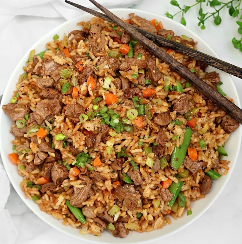 A bowl of fried rice with chunks of vegan meat, carrots, green beans, and green onions, garnished with herbs. Two chopsticks rest on the rim of the bowl.