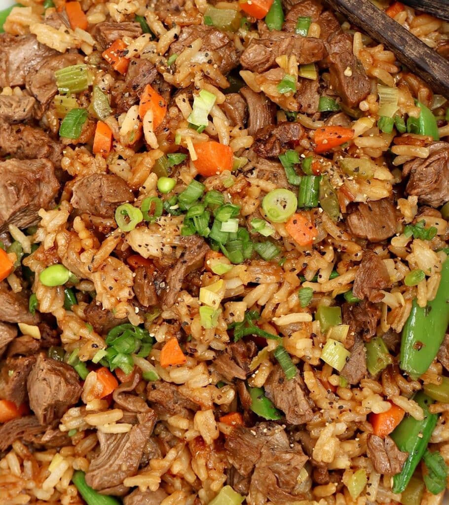 Close-up of fried rice with chunks of plant-based beef, diced carrots, peas, chopped green onions, and snap peas, garnished with black pepper and served with chopsticks.