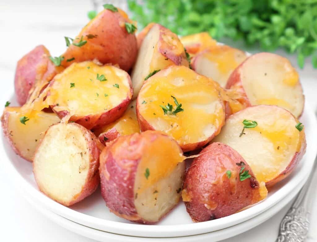 A plate of roasted red potatoes topped with melted cheddar cheese and sprinkled with chopped parsley, with a green leafy garnish in the background.