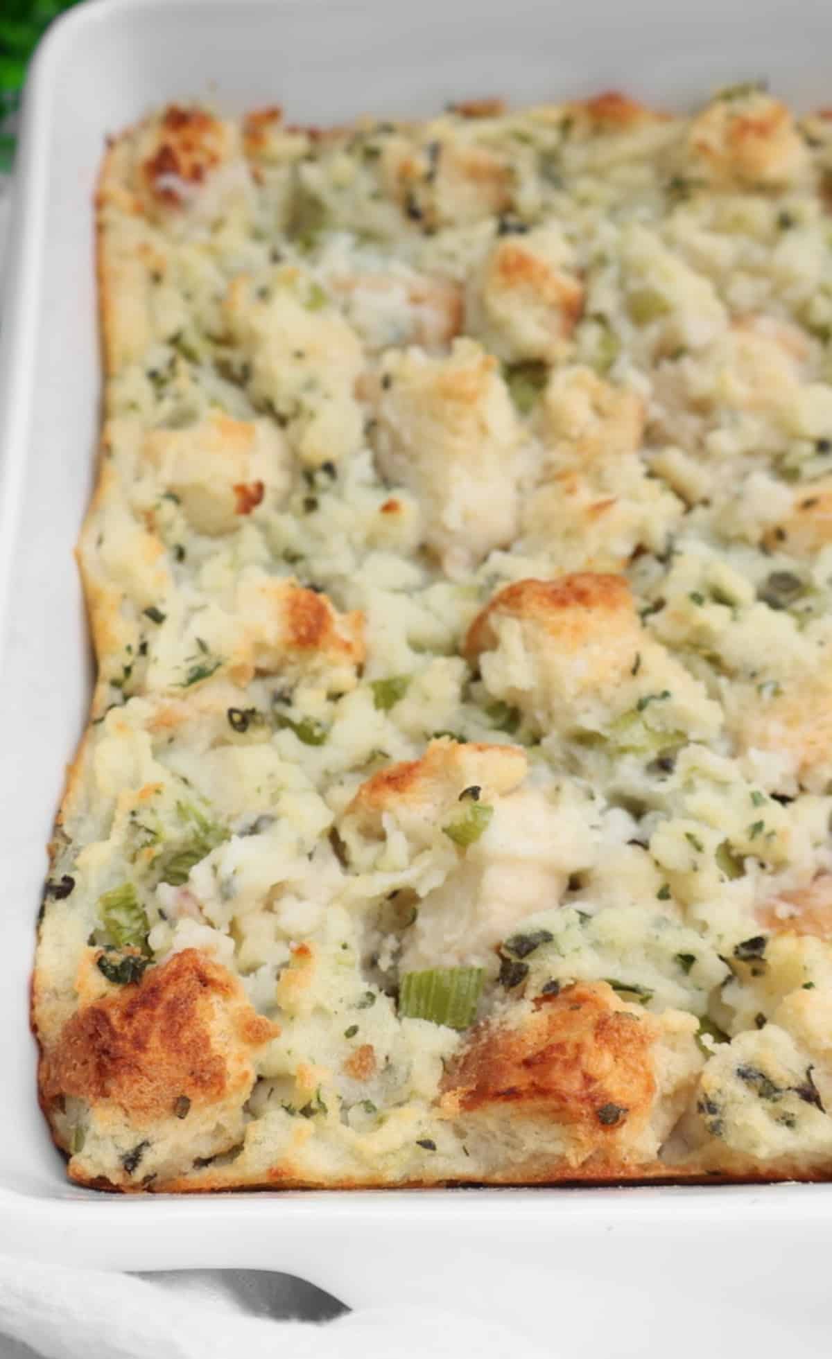 A close-up of a baked bread stuffing casserole in a white dish, featuring golden brown bread cubes mixed with herbs and celery.