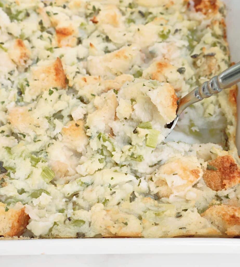A close-up of a baking dish filled with bread stuffing. The stuffing contains chunks of bread, celery pieces, and herbs. A metal serving spoon is scooping out a portion from the dish.