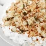 A close-up of a creamy dip topped with breadcrumbs and chopped pickles, served on a white plate. The dip appears thick and garnished for added texture and flavor.