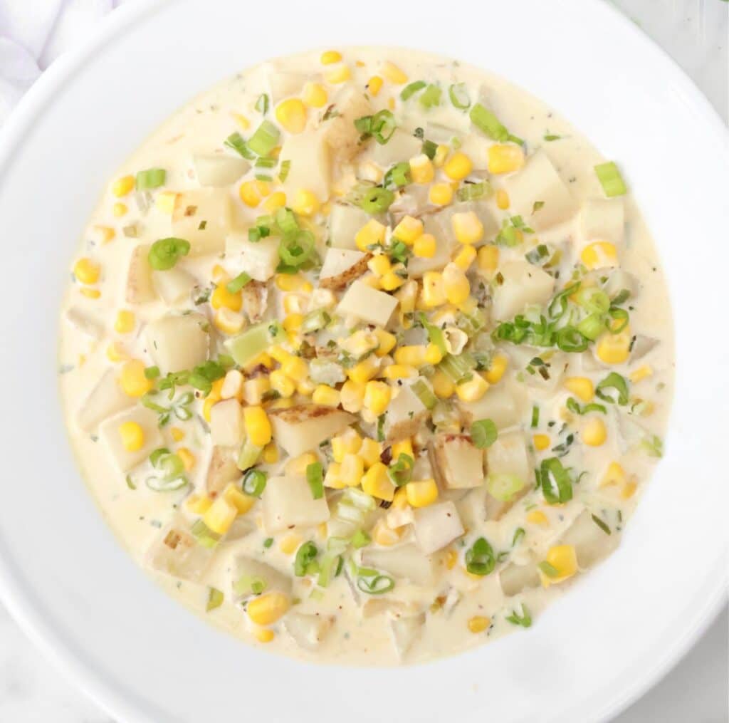 A bowl of creamy corn chowder with diced potatoes, corn kernels, sliced green onions, and herbs, served in a white dish on a marble surface.