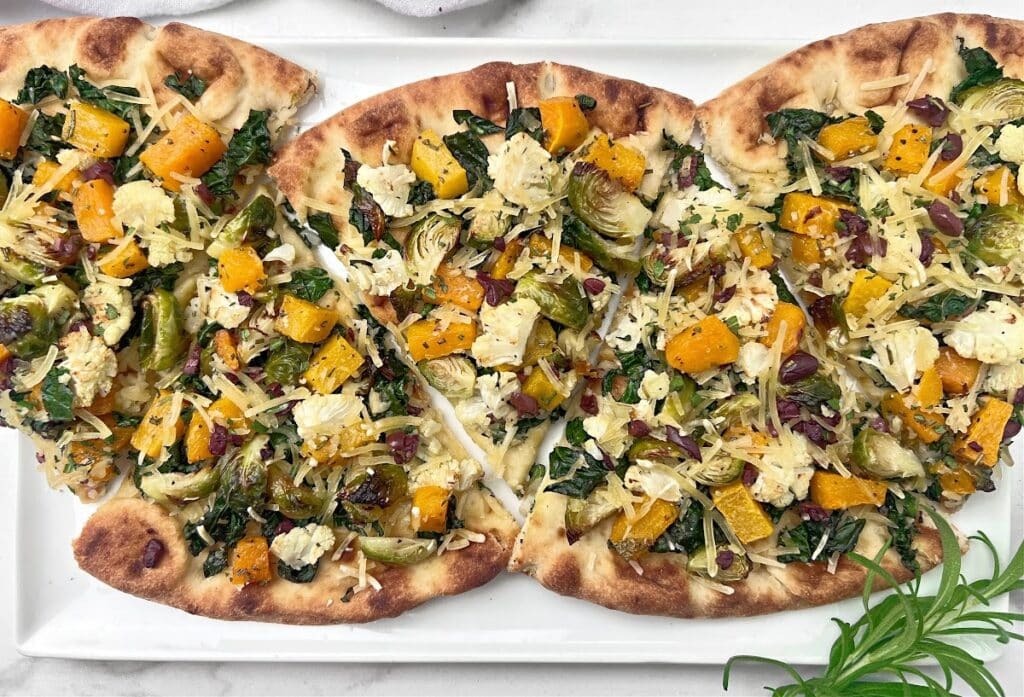 Three flatbread pizzas topped with roasted butternut squash, Brussels sprouts, kale, cheese, and herbs are arranged on a white platter, garnished with a fresh sprig of rosemary.